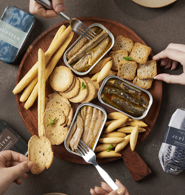 Tábua de madeira com conservas de peixe Jupiter em azeite biológico, acompanhadas por tostas, grissinos e mãos a servir, criando um momento de partilha gourmet.